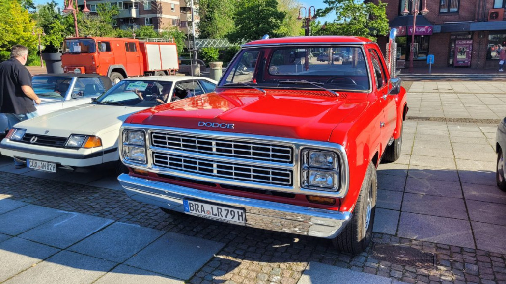 Ein leuchtend roter Dodge-Oldtimer-Truck steht auf einer gepflasterten Fläche, im Hintergrund sind Autos und Menschen zu sehen. Der Truck hat einen glänzenden Kühlergrill und ein klassisches Design.